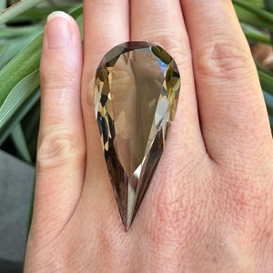 May include: A large, pear-shaped faceted gemstone with a translucent, smoky brown color. The stone features numerous triangular and trapezoidal facets that create a reflective, geometric surface across its elongated, pointed teardrop silhouette.