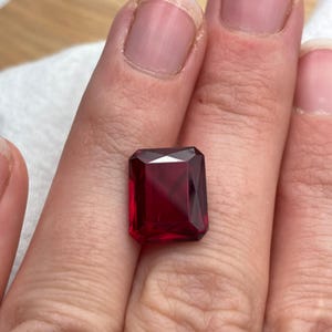 May include: A faceted, rectangular-cut, deep red gemstone. The gem is held in a hand, showcasing its clarity and color. The gemstone's facets reflect light, creating a brilliant appearance. This gem could be used in jewelry.