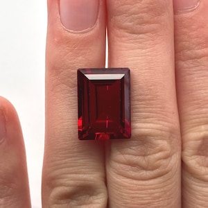 Peut inclure: Une pierre précieuse rouge profond avec une taille émeraude rectangulaire, présentant des facettes nettes et précises qui reflètent la lumière. La pierre affiche une teinte cramoisie riche et saturée sur tout son corps transparent.