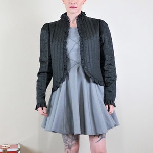 May include: A grey dress with a flared skirt and a black quilted jacket with ruffled trim. The outfit is paired with black velvet ankle boots. A red and white trunk is on the left.