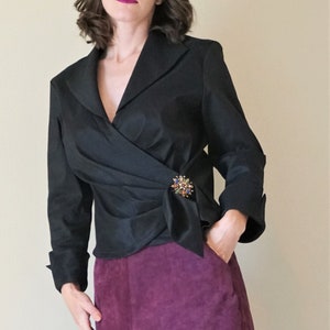 May include: A black wrap-style top with long sleeves and a decorative brooch. The top is paired with a knee-length, deep purple suede skirt. The outfit is a classic, elegant style.