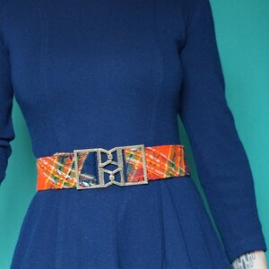 May include: A navy blue dress with a long sleeve and a fitted bodice. The dress has a flared skirt and is cinched at the waist with a colorful patterned belt with a gold buckle.