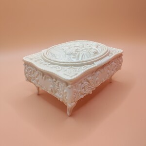Puede incluir: Una caja de joyería blanca adornada con un diseño floral y tres patas. La caja tiene un óvalo elevado en el centro con un diseño floral.