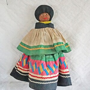 May include: A handmade fabric doll with a black head, brown eyes, and a yellow beaded necklace. The doll is wearing a beige skirt with green and yellow trim, a green skirt with a pink and blue patchwork design, and a black skirt with white trim.