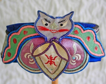 Può includere: Un cappello vintage ornato con una faccia di gatto in cima. Il cappello presenta una base blu con dettagli ricamati in verde, rosa e viola. Un ciondolo con un simbolo rosso è appeso sotto la faccia del gatto. Il cappello è probabilmente di origine asiatica.
