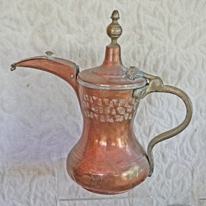 May include: A copper and brass traditional Arabic coffee pot with a decorative embossed design. The pot has a hinged lid and a spout with a small handle. The handle is made of brass and is attached to the pot with a hinge.