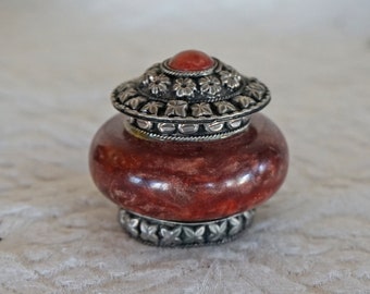May include: A small, round, red and brown stone box with a silver lid. The lid is decorated with a silver band and a red stone in the center.