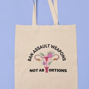 May include: A beige tote bag with a black graphic that says "Ban Assault Weapons Not Abortions" with a stylized illustration of a uterus with flowers.