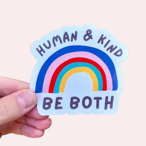 May include: A rainbow sticker with the text "Human & Kind Be Both" in black lettering. The sticker is on a white background.