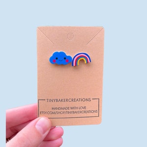 May include: A pair of handmade earrings featuring a blue cloud with a smiling face and a rainbow. The earrings are displayed on a brown card with the text "TINYBAKERCREATIONS" and "HANDMADE WITH LOVE" and the website address "ETSY.COM/SHOP/TINYBAKERCREATIONS".