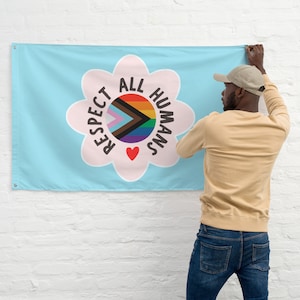 May include: A light blue flag with a pink flower design. The flower has the words "Respect All Humans" written around it. Inside the flower is a rainbow heart with a black and brown arrow pointing to the left.