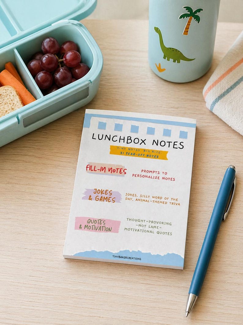 Może przedstawiać: Jasnoniebieski pojemnik na lunch z winogronami, marchewką i chlebem, butelka na wodę z motywem dinozaura i notatnik z tekstem "LUNCHBOX NOTES". Niebieski długopis spoczywa na drewnianej powierzchni. Notatnik zawiera podpowiedzi do notatek, żart&oacute;w i cytat&oacute;w.