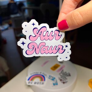 Peut inclure: Un autocollant rose et bleu avec le texte "Aur Naur" dans une police r&eacute;tro. L'autocollant a un fond blanc et est d&eacute;cor&eacute; de petites &eacute;toiles blanches.