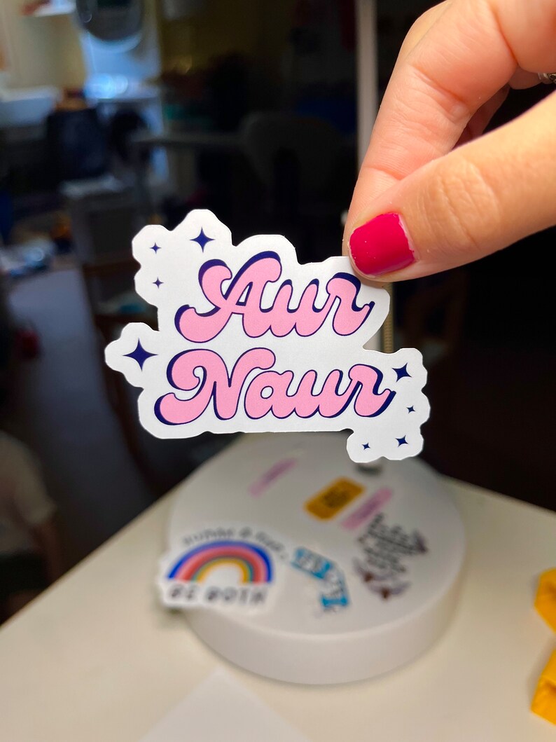 Peut inclure: Un autocollant rose et bleu avec le texte "Aur Naur" dans une police r&eacute;tro. L'autocollant a un fond blanc et est d&eacute;cor&eacute; de petites &eacute;toiles blanches.
