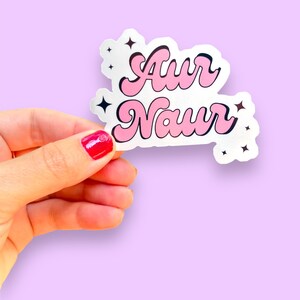 Peut inclure: Un autocollant rose et blanc avec le texte "Aur Naur" dans une police r&eacute;tro. L'autocollant a de petites &eacute;toiles noires autour du texte.