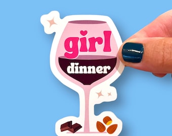 May include: A sticker featuring a wine glass design with the words "girl dinner" in pink and white. The glass is filled with a dark red liquid. Chocolate pieces and almonds are at the bottom of the sticker. The sticker is held by a hand.