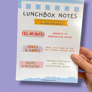Może przedstawiać: Notatnik zatytułowany "LUNCHBOX NOTES" z odrywanymi stronami. Okładka zawiera tekst w r&oacute;żnych kolorach, w tym "FILL-IN NOTES", "JOKES & GAMES" i "QUOTES & MOTIVATION". Notatnik jest trzymany przez osobę.