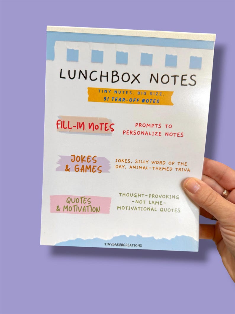 Może przedstawiać: Notatnik zatytułowany "LUNCHBOX NOTES" z odrywanymi stronami. Okładka zawiera tekst w r&oacute;żnych kolorach, w tym "FILL-IN NOTES", "JOKES & GAMES" i "QUOTES & MOTIVATION". Notatnik jest trzymany przez osobę.
