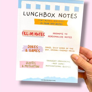 Może przedstawiać: Biały notatnik Lunchbox Notes z niebieskimi i ż&oacute;łtymi akcentami trzymany w dłoni. Notatnik zawiera tekst, w tym "Lunchbox Notes", "Fill-in Notes", "Jokes & Games" i "Quotes & Motivation". Notatnik zawiera 51 karteczek.