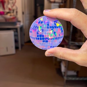 May include: A holographic disco ball sticker with a purple, blue, and pink iridescent finish. The sticker is round and has a mirrored surface.