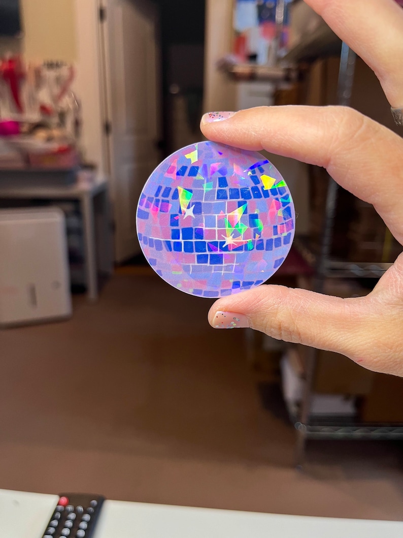 May include: A holographic disco ball sticker with a purple, blue, and pink iridescent finish. The sticker is round and has a mirrored surface.
