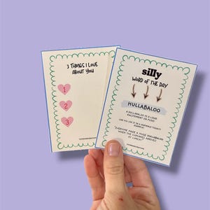 Może przedstawiać: Dwie karty trzymane w dłoni, z zabawnymi tekstami i wzorami. Jedna karta głosi "3 THINGS I LOVE ABOUT YOU" z ikonami serca. Druga karta m&oacute;wi "silly WORD OF THE DAY" ze słowem "HULLABALOO" i jego definicją.