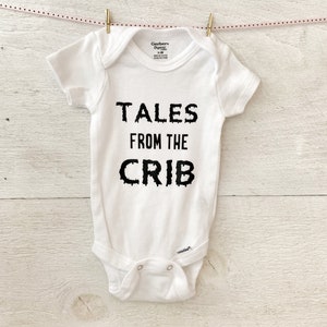 K&ouml;nnte beinhalten: Wei&szlig;er Baby-Strampler mit schwarzem Text, der "Tales from the Crib" lautet.