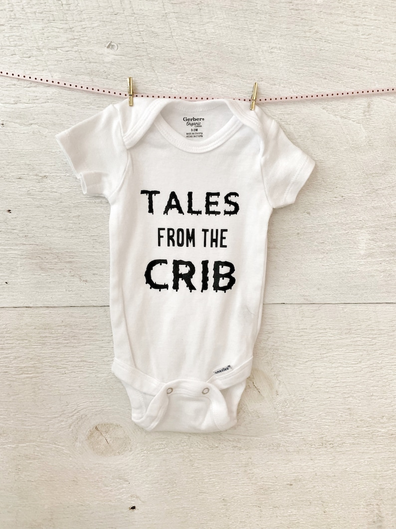 K&ouml;nnte beinhalten: Wei&szlig;er Baby-Strampler mit schwarzem Text, der "Tales from the Crib" lautet.