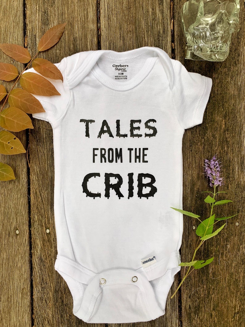 K&ouml;nnte beinhalten: Wei&szlig;er Baby-Strampler mit schwarzem, tropfendem Text, der "Tales from the Crib" lautet.