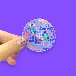 May include: A clear iridescent disco ball sticker with a holographic finish. The sticker has a grid pattern and is decorated with small, colorful, iridescent pieces.