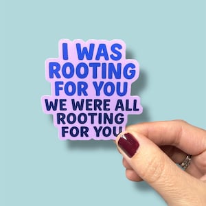 May include: A sticker with the text "I WAS ROOTING FOR YOU WE WERE ALL ROOTING FOR YOU" in blue font on a light purple background. The sticker is held by a hand with a maroon nail polish.
