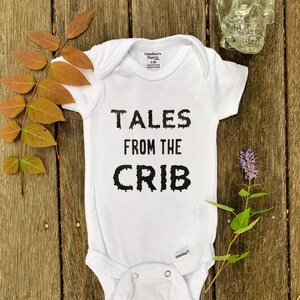 K&ouml;nnte beinhalten: Wei&szlig;er Baby-Strampler mit schwarzem Text, der "Tales from the Crib" lautet.