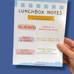 Może przedstawiać: Biała książeczka Lunchbox Notes z niebiesko-białym wzorem z rozdartym papierem. Okładka zawiera tytuł "LUNCHBOX NOTES" oraz sekcje na notatki do wypełnienia, żarty i gry oraz cytaty i motywację. Książeczka zawiera 51 karteczek do oderwania.