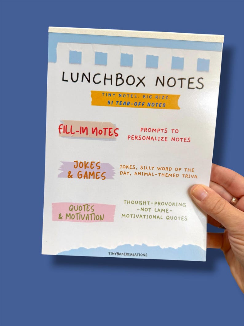 Może przedstawiać: Biała książeczka Lunchbox Notes z niebiesko-białym wzorem z rozdartym papierem. Okładka zawiera tytuł "LUNCHBOX NOTES" oraz sekcje na notatki do wypełnienia, żarty i gry oraz cytaty i motywację. Książeczka zawiera 51 karteczek do oderwania.