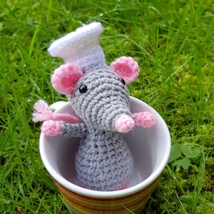 Puede incluir: Una rata de ganchillo hecha a mano con un gorro de chef, sentada dentro de una taza a rayas. La rata es gris con orejas, patas y nariz rosas. La taza está sobre hierba verde.