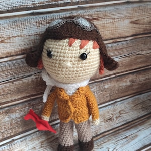 Op de afbeelding: Handgemaakte gehaakte pop van een piloot, met een beige gezicht, bruin haar en een bruine pilotenmuts met een bril. De pop draagt een mosterdgele jas, beige broek en een witte sjaal. Ze houdt een rood papieren vliegtuig vast.