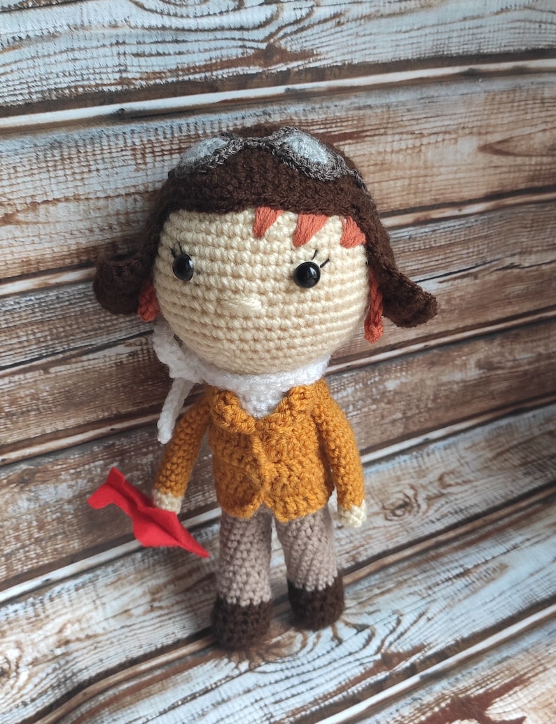 Op de afbeelding: Handgemaakte gehaakte pop van een piloot, met een beige gezicht, bruin haar en een bruine pilotenmuts met een bril. De pop draagt een mosterdgele jas, beige broek en een witte sjaal. Ze houdt een rood papieren vliegtuig vast.