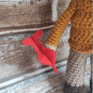 Op de afbeelding: Gehaakte pop met een mosterdgele jas, witte kraag en grijze broek. De pop heeft een rood vilten accessoire in haar hand. De pop is handgemaakt en heeft een grillig ontwerp.