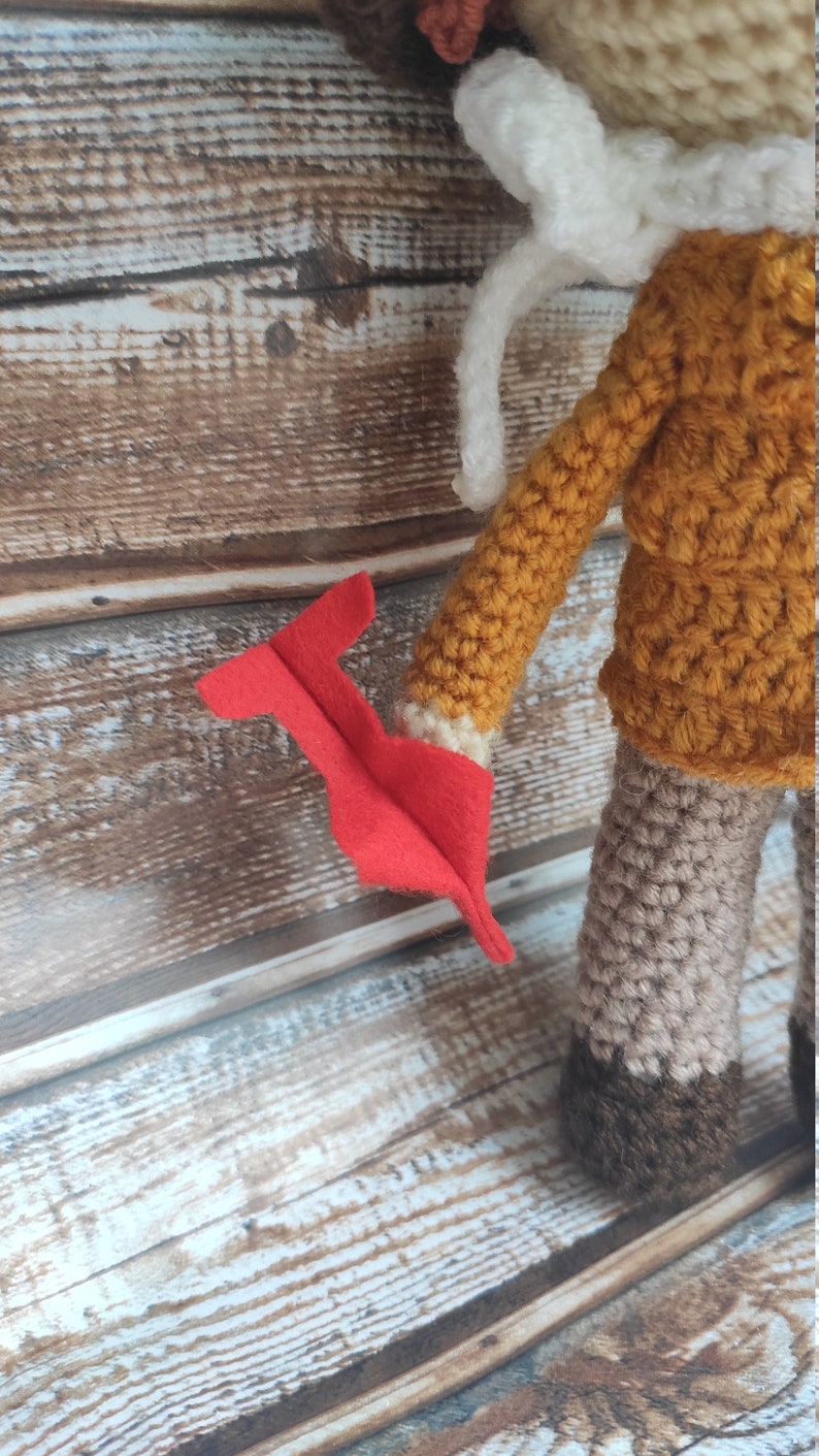 Op de afbeelding: Gehaakte pop met een mosterdgele jas, witte kraag en grijze broek. De pop heeft een rood vilten accessoire in haar hand. De pop is handgemaakt en heeft een grillig ontwerp.