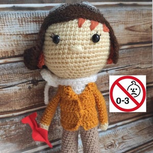Op de afbeelding: Handgemaakte gehaakte pop verkleed als piloot, met een beige gezicht, bruin haar en een oranje jas. De pop houdt een rood papieren vliegtuig vast. De afbeelding bevat een waarschuwingsbord dat aangeeft dat het speelgoed niet geschikt is voor kinderen van 0-3 jaar.