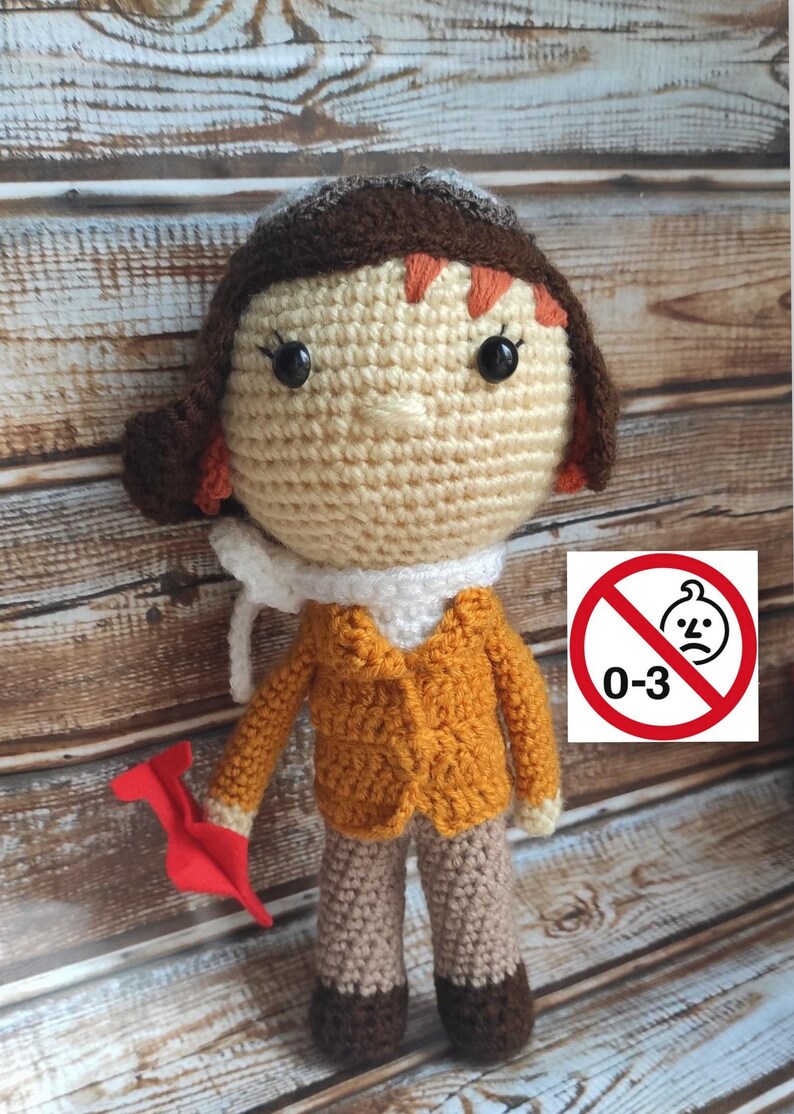 Op de afbeelding: Handgemaakte gehaakte pop verkleed als piloot, met een beige gezicht, bruin haar en een oranje jas. De pop houdt een rood papieren vliegtuig vast. De afbeelding bevat een waarschuwingsbord dat aangeeft dat het speelgoed niet geschikt is voor kinderen van 0-3 jaar.