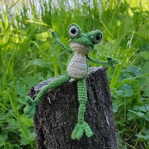 May include: A handmade crochet frog toy featuring a bright green body with a cream-colored belly, large black plastic eyes, and long limbs. The frog is posed with one arm raised and one leg extended, showcasing detailed stitching and yarn texture.
