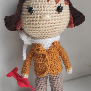 Op de afbeelding: Gehaakte pop van een piloot met een bruine pilotenmuts, oranje jas en beige broek. De pop heeft een witte sjaal en houdt een rood vliegtuig vast. Het haar van de pop is roodbruin.