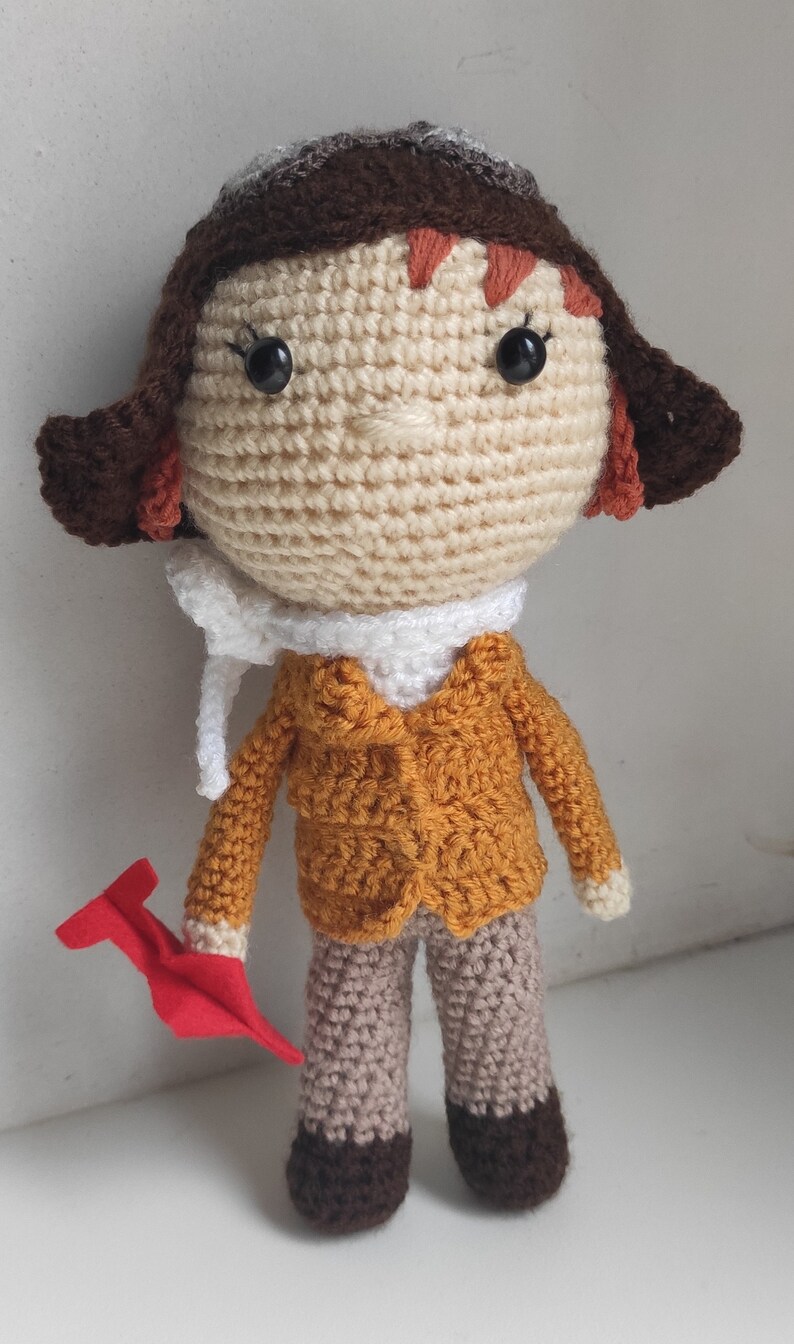 Op de afbeelding: Gehaakte pop van een piloot met een bruine pilotenmuts, oranje jas en beige broek. De pop heeft een witte sjaal en houdt een rood vliegtuig vast. Het haar van de pop is roodbruin.