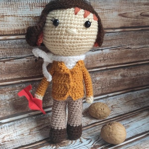 Op de afbeelding: Gehaakte pop van een piloot, met een bruine pilotenmuts, oranje jas en beige broek. De pop houdt een rood papieren vliegtuig vast. De pop is ongeveer 25 cm hoog. Op de achtergrond liggen drie walnoten.
