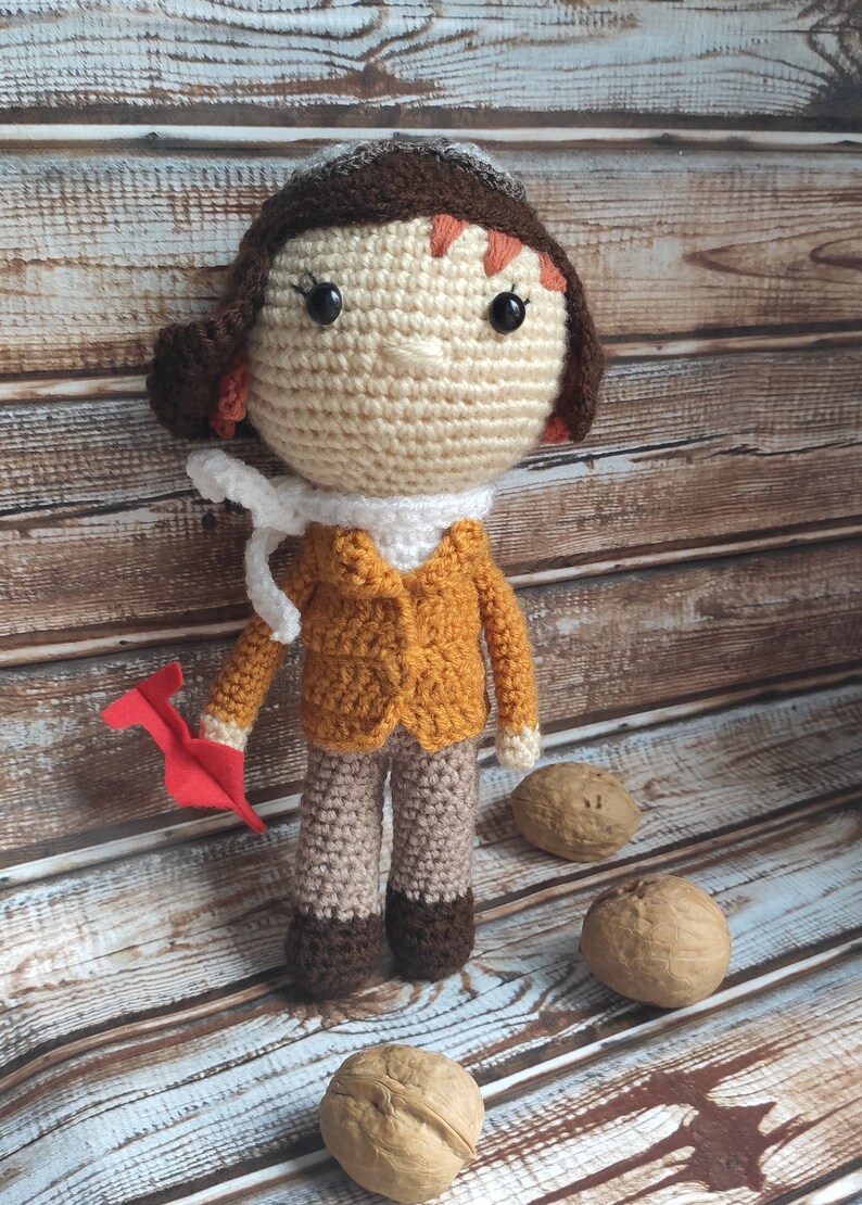 Op de afbeelding: Gehaakte pop van een piloot, met een bruine pilotenmuts, oranje jas en beige broek. De pop houdt een rood papieren vliegtuig vast. De pop is ongeveer 25 cm hoog. Op de achtergrond liggen drie walnoten.