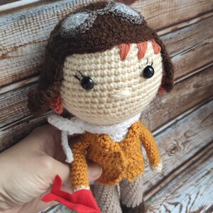 Op de afbeelding: Gehaakte pop van een vrouwelijke piloot, met een beige gezicht, bruin haar en een mosterdkleurig jasje. De pop draagt een bril en een witte sjaal en houdt een rood vliegtuig vast. Een charmant handgemaakt item.
