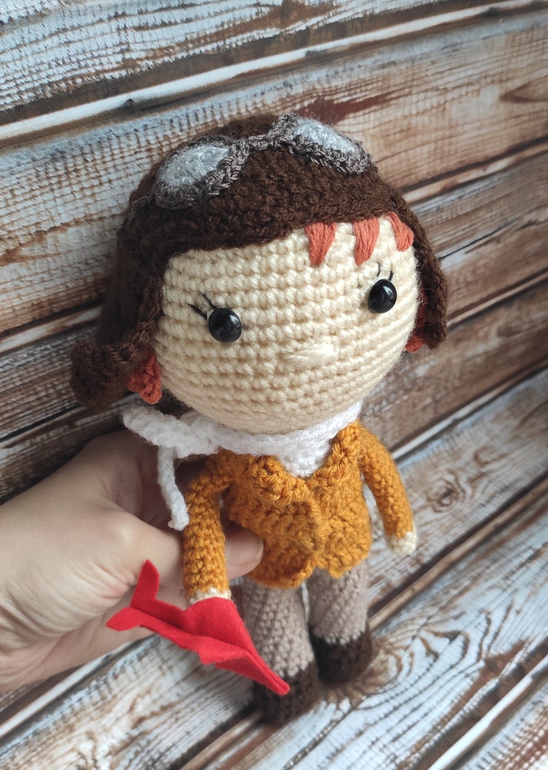 Op de afbeelding: Gehaakte pop van een vrouwelijke piloot, met een beige gezicht, bruin haar en een mosterdkleurig jasje. De pop draagt een bril en een witte sjaal en houdt een rood vliegtuig vast. Een charmant handgemaakt item.