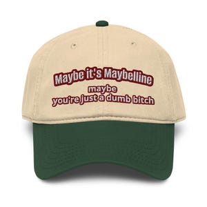 May include: A beige baseball cap with a dark green brim and button. The cap features embroidered text in a burgundy color that reads "Maybe it's Maybelline, maybe you're just a dumb bitch."
