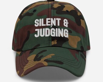 Könnte beinhalten: Eine Baseballkappe mit grünem, braunem und schwarzem Tarnmuster. Auf der Vorderseite der Kappe befindet sich der weiß gestickte Text "SILENT & JUDGING" in einer fetten serifenlosen Schrift. Die Kappe hat einen gebogenen Schirm und eine strukturierte Krone.